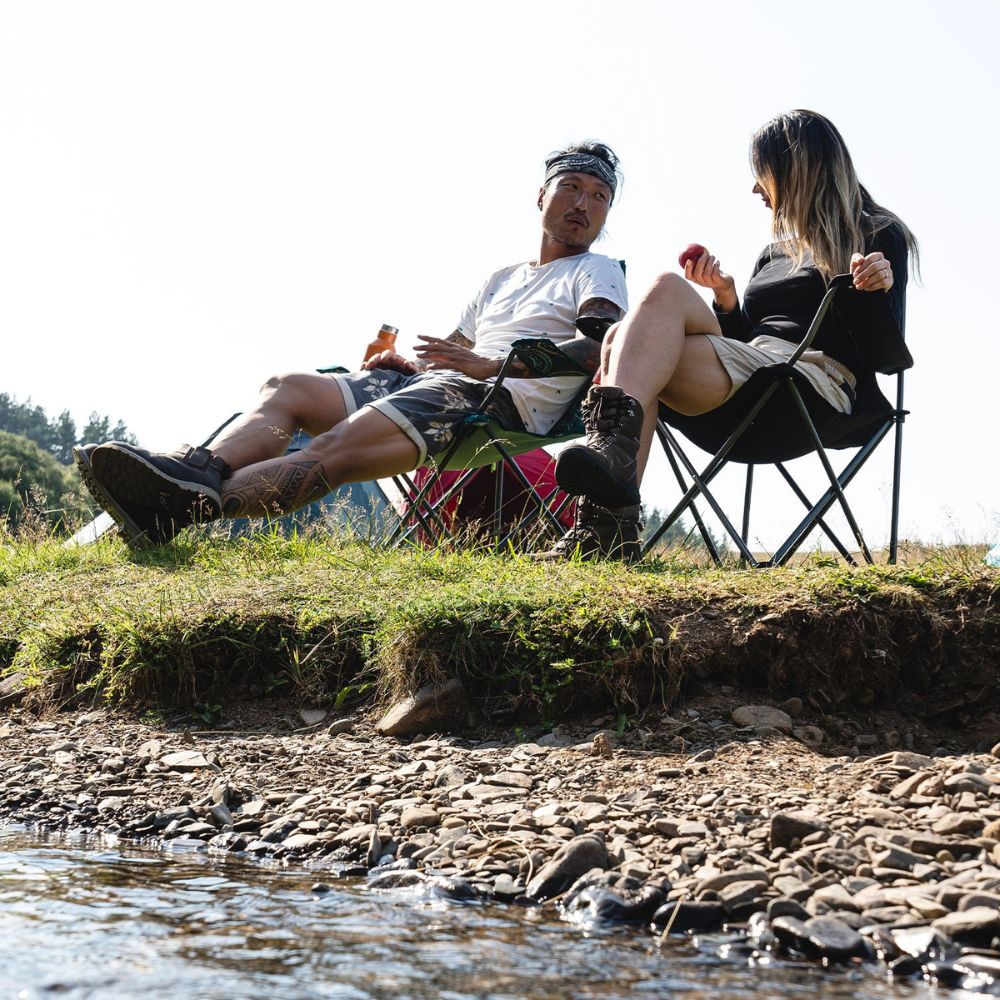 Two campers relaxing by a riverside, sitting on black Edinburgh camping chairs.