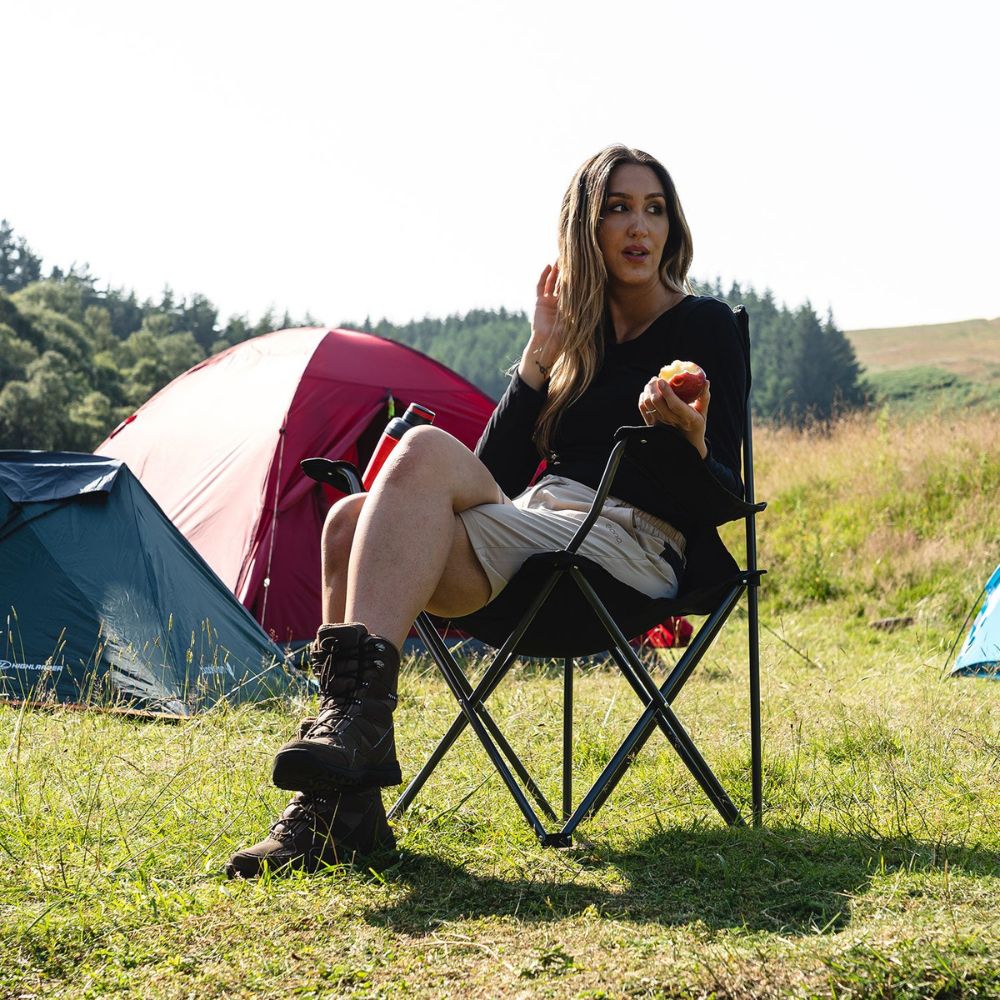 A camper holding an apple while seated on a black Edinburgh camping chair near a tent.
