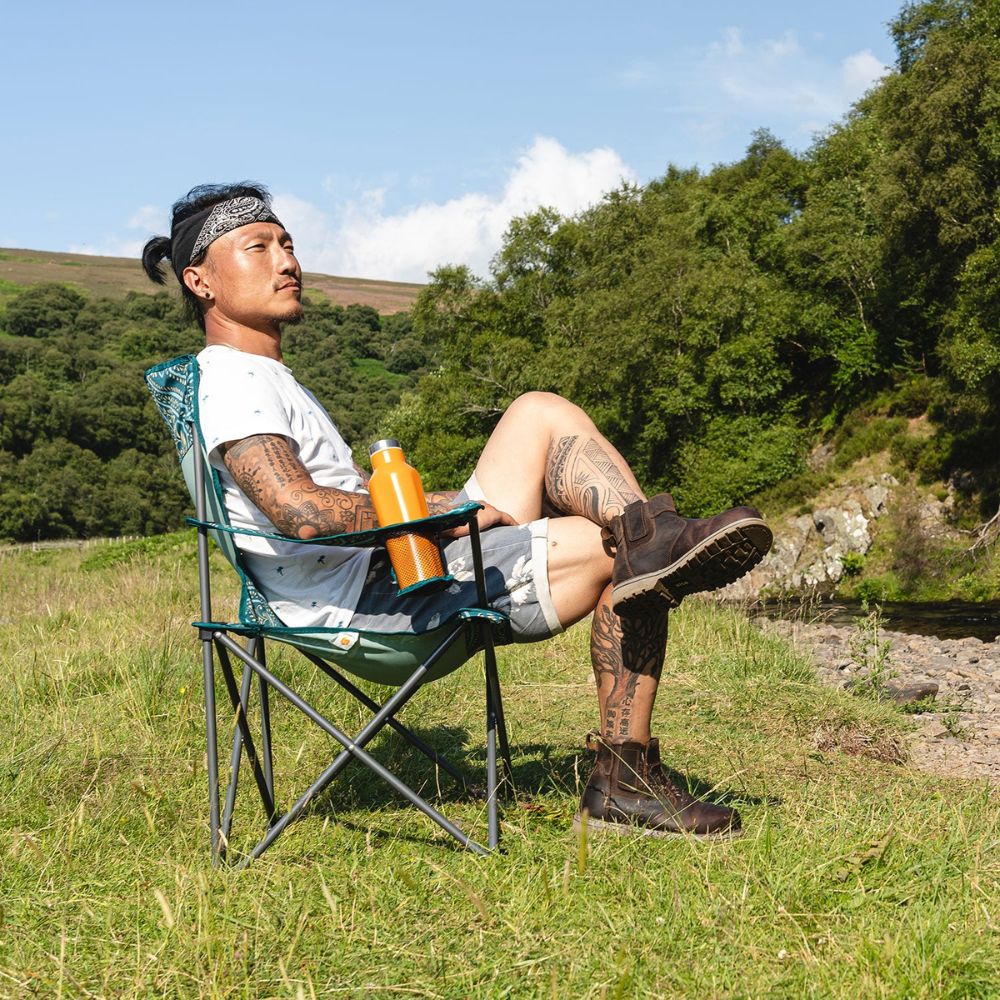 A camper sitting on a black Edinburgh camping chair, enjoying the outdoors by a river.