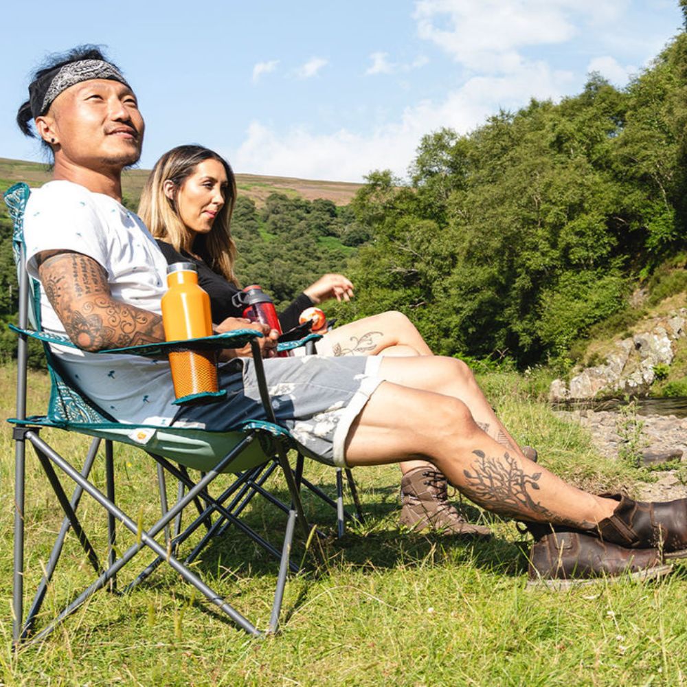 Two campers seated on black Edinburgh camping chairs, enjoying a riverside view.