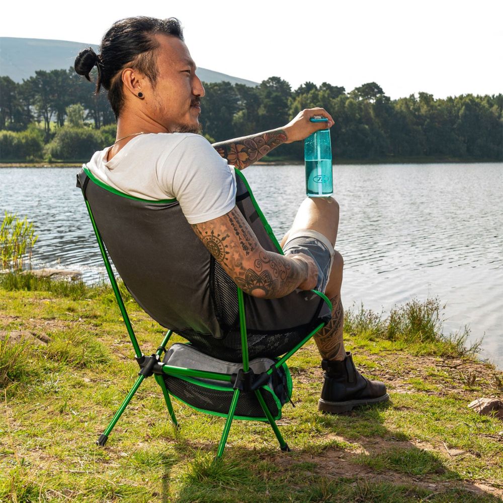 A person relaxes in a Highlander Ayr Folding Camping Chair on the shore of a lake.