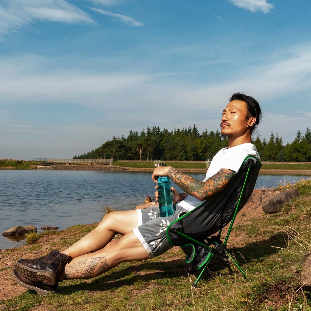 A person relaxing in a Highlander Ayr Folding Camping Chair by a lake, enjoying the scenery.