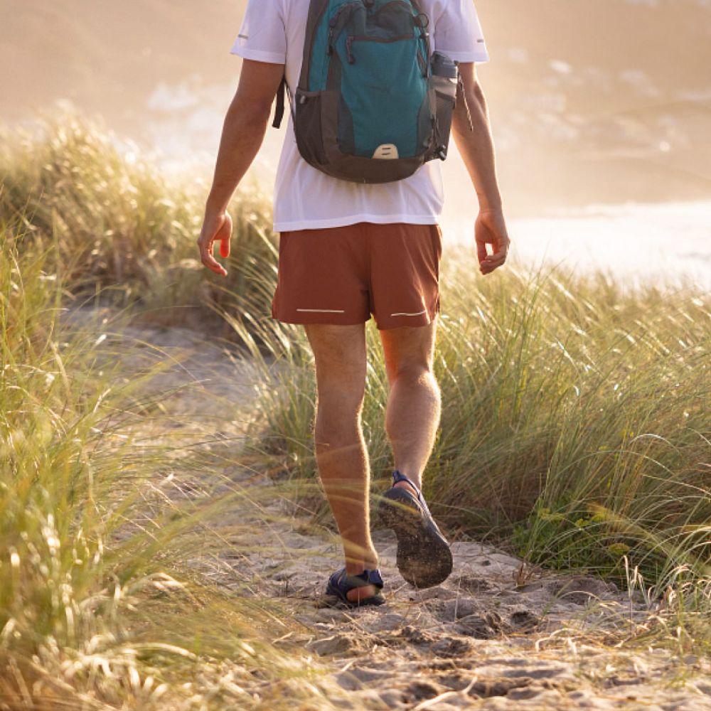 A man wearing a pair of Keen Newport H2 sandals walks on a sandy trail in the sunshine.