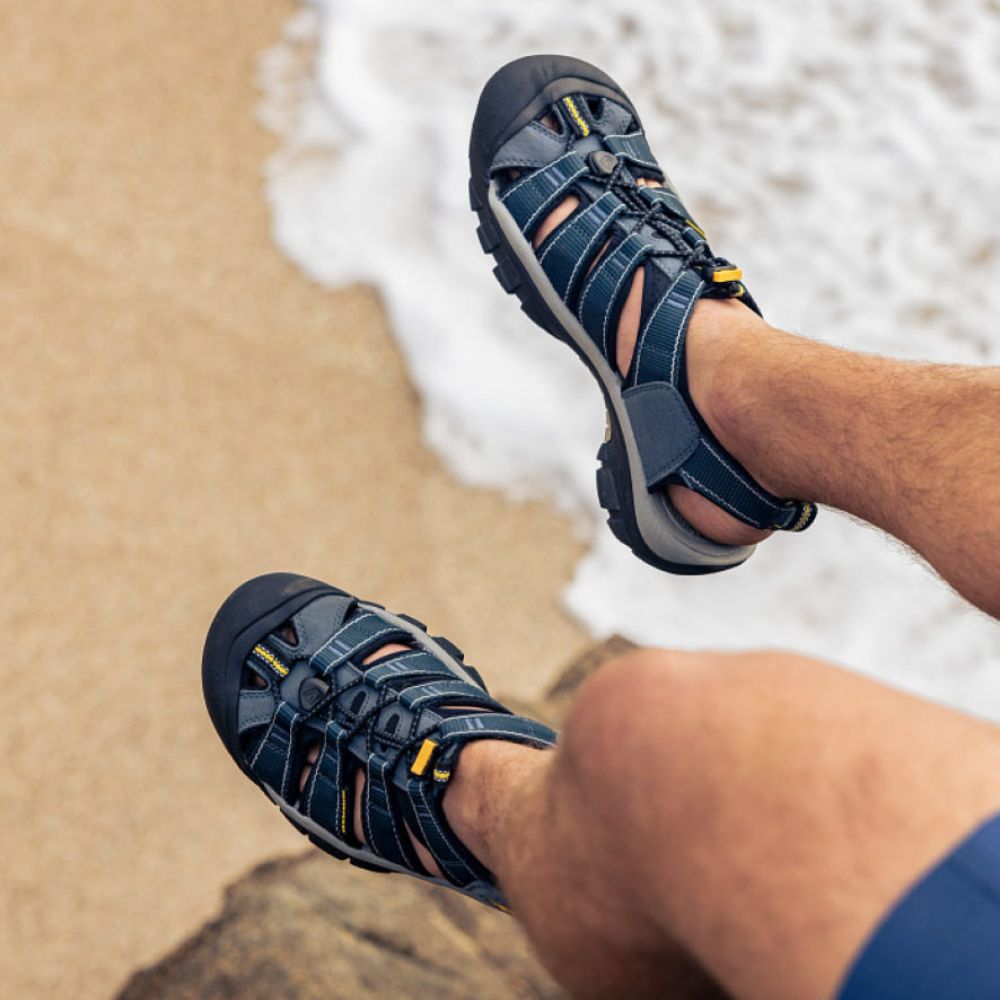 A person wearing a pair of KEEN Men's Newport H2 sandals in blue and yellow, sitting on a rock.