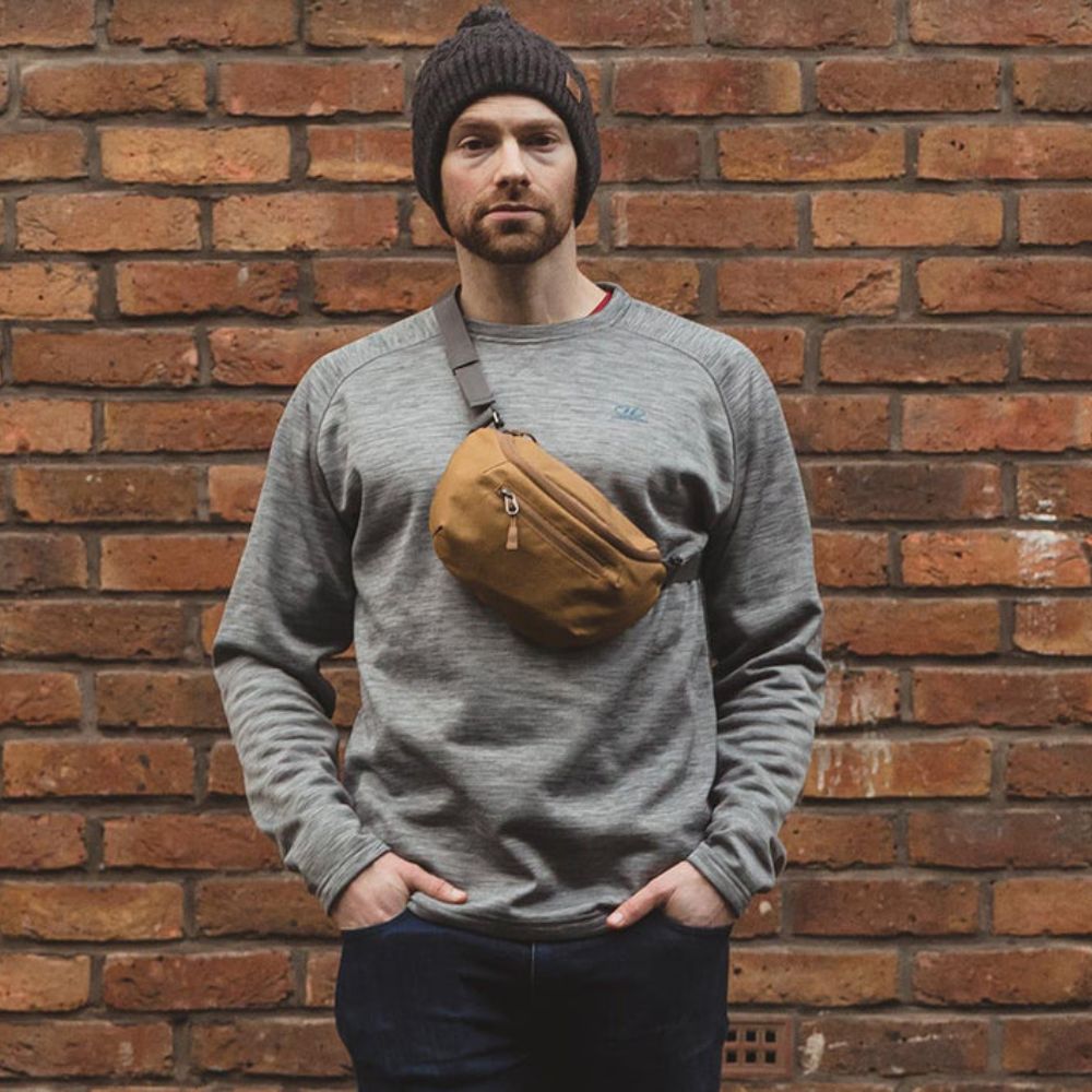 Man standing against a brick wall wearing a grey sweatshirt and a mustard Targa bum bag.